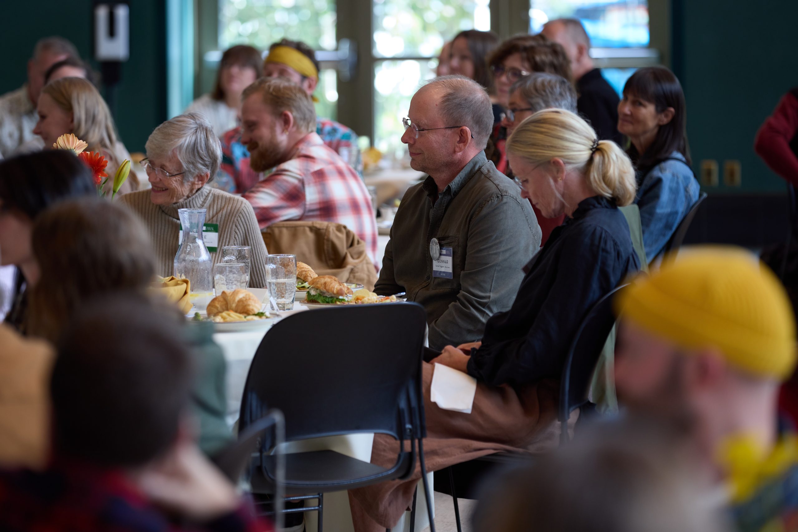 This image shows a group of artists and guests enjoying lunch and listening to someone speaking out of the picture