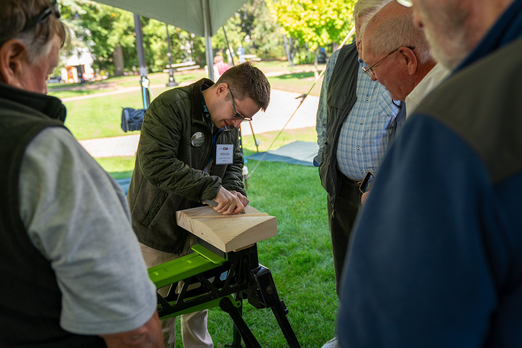 This image shows artist Spencer Tinkham carving a block of wood in front of four men watching him