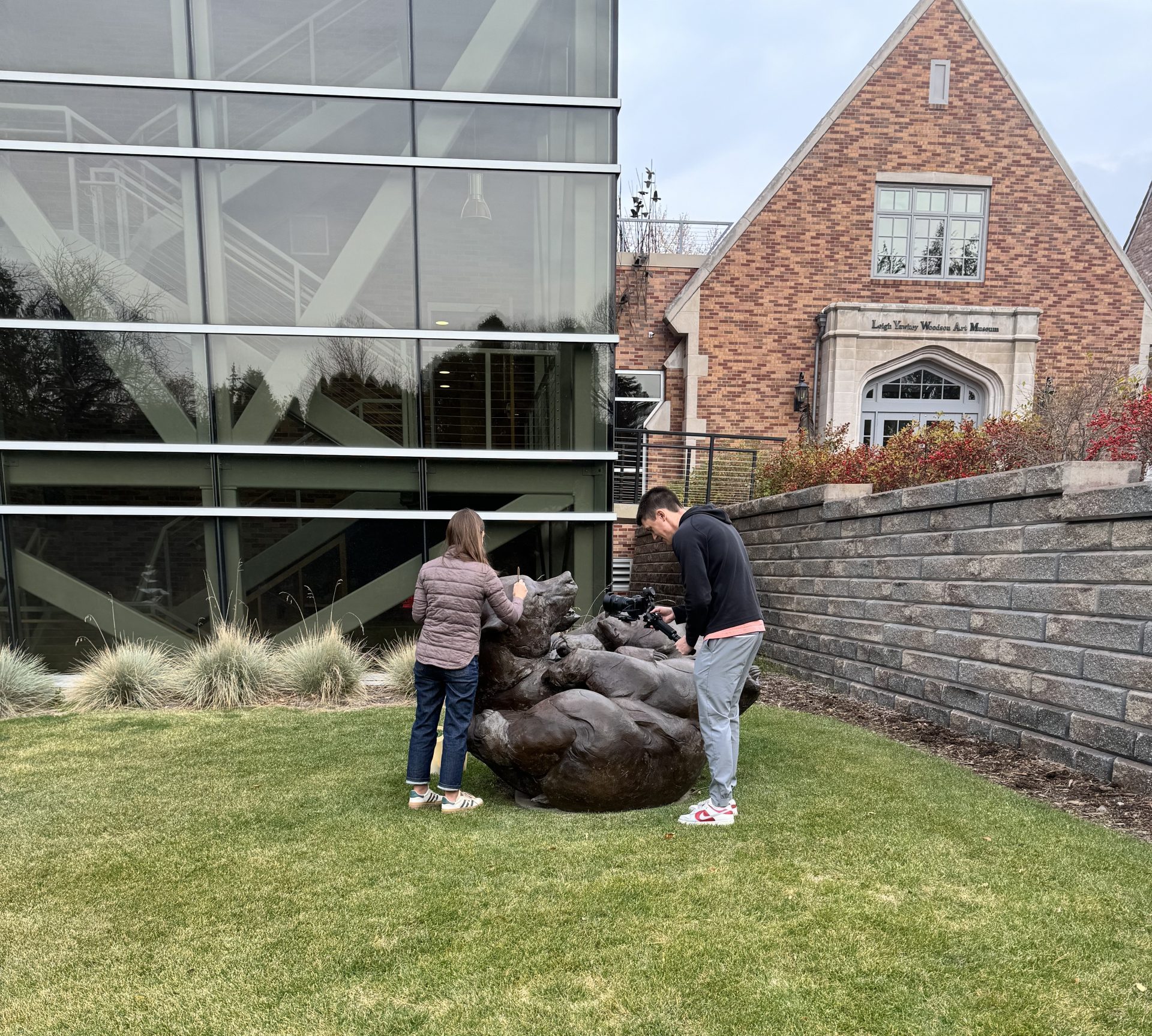 Shannon is outside cleaning a large bronze sculpture of two large bears playing. There is a man with a video camera recording her.