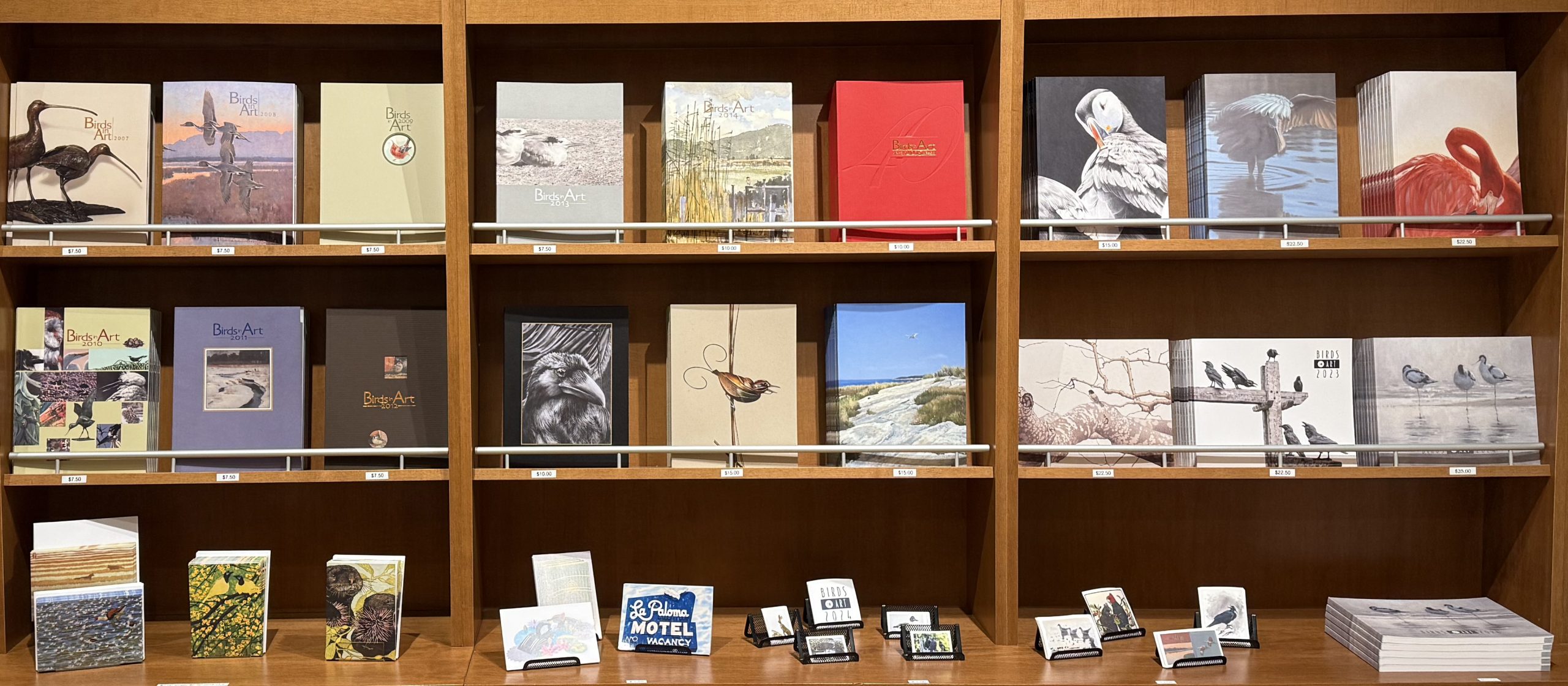 catalogues of birds in art sit in a rack nine across and two tall on the lower shelf there are artworks on notecards, postcards, and stickers