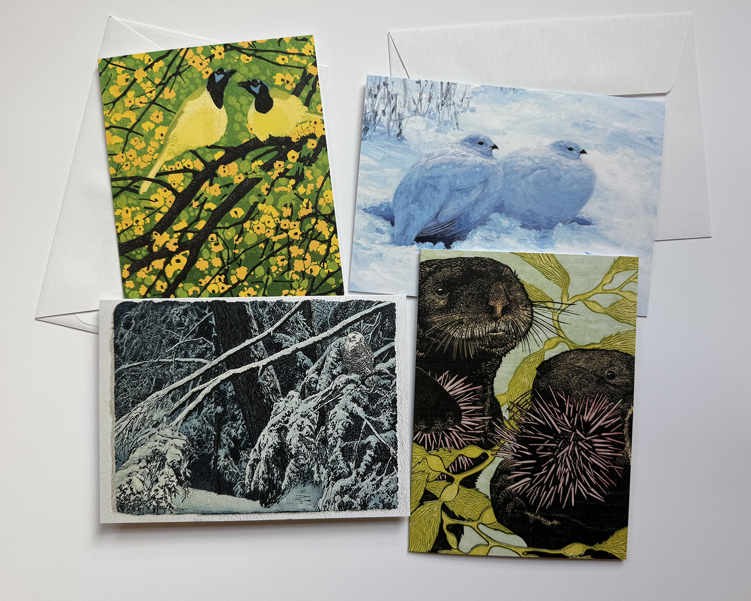 four notecards, one with two yellow birds sitting in a tree with yellow flowers, one is two puffy birds sitting in snow, the next is an owl on a snowy branch, and the last is two otters with sea weed 